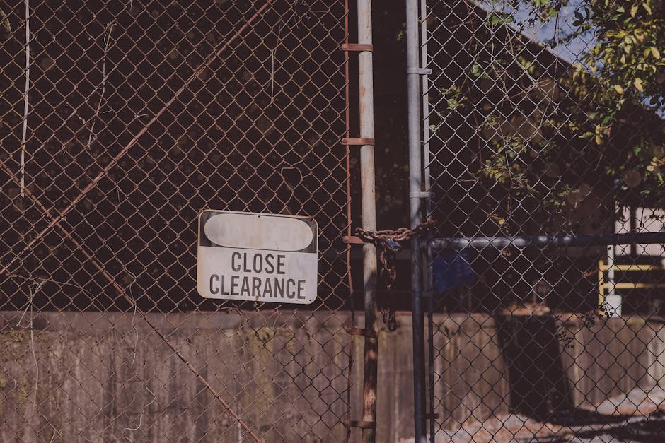 A close-up view of a weathered metal gate composed of a diamond-shaped wire mesh, secured with a rusty chain and padlock. The gate is painted in a dark hue, with visible signs of rust and corrosion, p